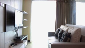 A modern living room with a flat-screen TV mounted on the wall, a sofa with patterned cushions, and a window with curtains. - Brand New 2BR @ Tamansari La Grande Apartment Bandung (Bandung)