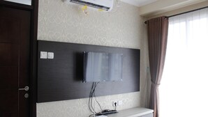 A hotel room with a mounted flat-screen TV, a wooden desk with a remote control, and a wall-mounted air conditioner. - Elegant 2BR Gateway Pasteur Apartment near Exit Toll Pasteur (Cimahi)