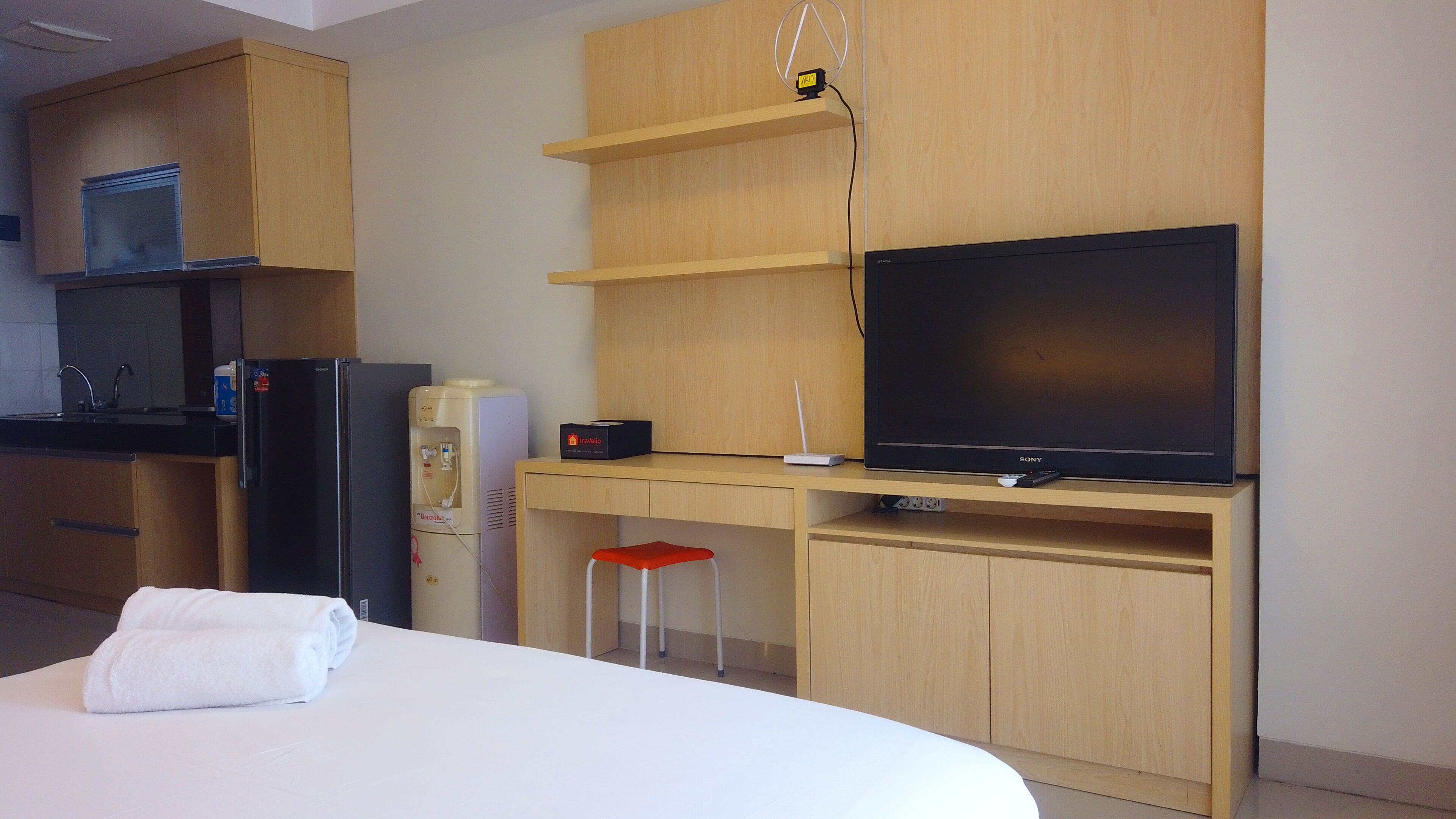Habitación de hotel con una cama, televisor, escritorio con computadora, dispensador de agua y un taburete pequeño.