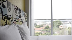 A hotel room with a bed, a large window with a view of buildings and greenery, and a patterned headboard. - Trendy 1BR near ITB @ Dago Suites Apartment (Bandung)