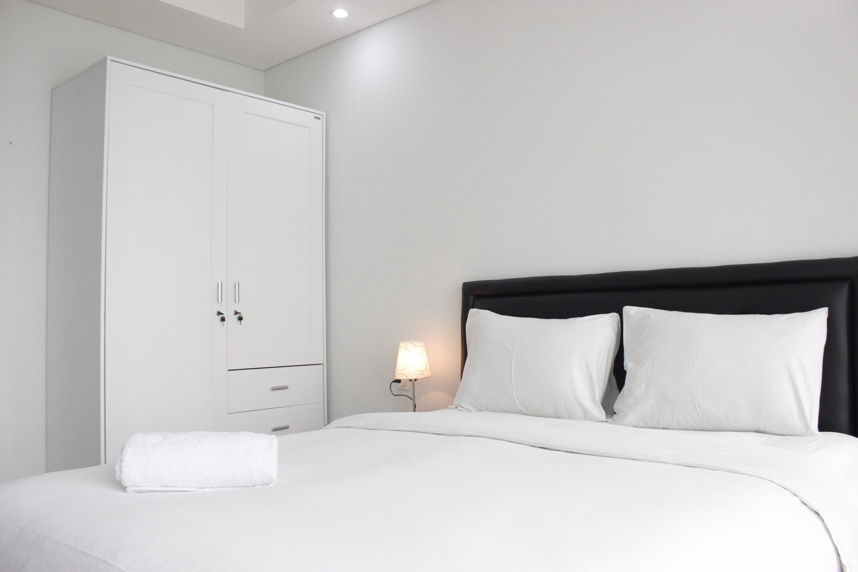 Una cama bien hecha con sábanas y almohadas blancas, un armario blanco y una lámpara de noche.