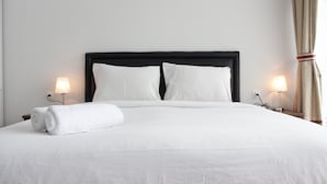A neatly made bed with white linens, two pillows, and a rolled towel on the side. There are two bedside lamps and a curtain on the right. - Strategic Studio at Green Kosambi Bandung Apartment (Bandung)