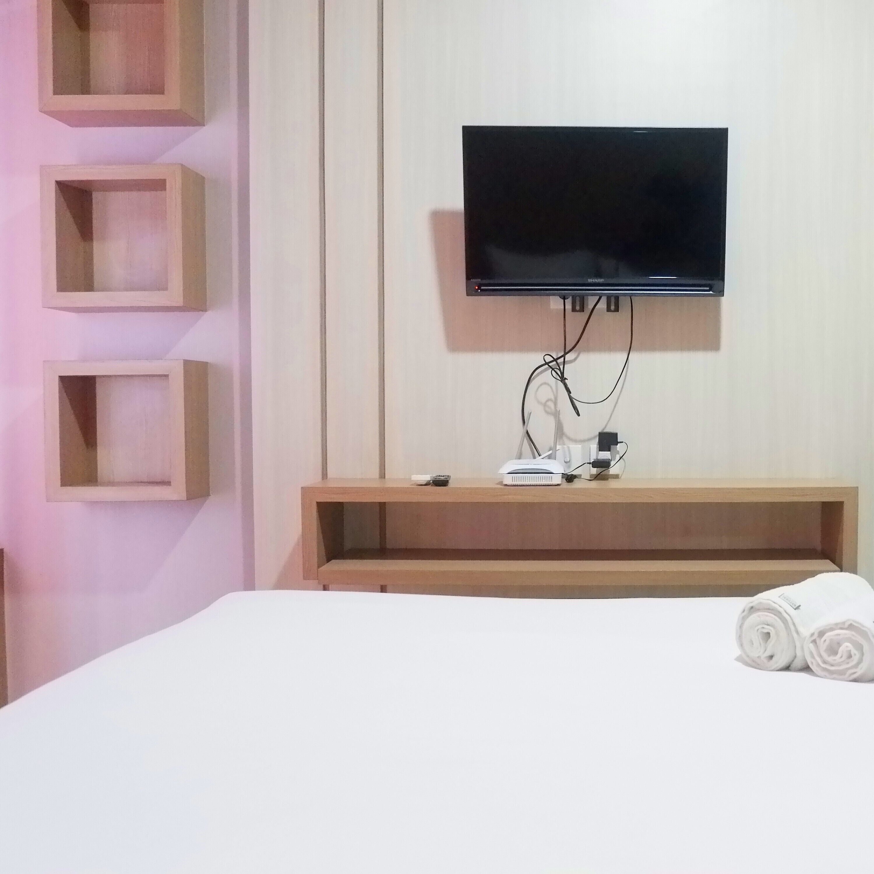 A hotel room with a flat-screen TV mounted on the wall, a wooden TV stand with a remote, and a neatly made bed with rolled towels.