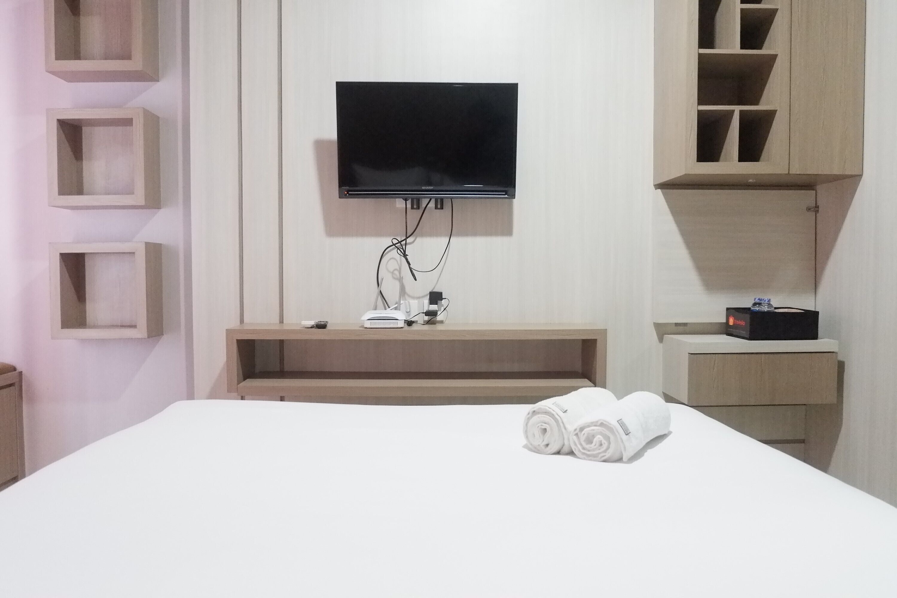 A hotel room with a flat-screen TV mounted on the wall, a bedside table with a hairdryer, and a neatly made bed with rolled towels.