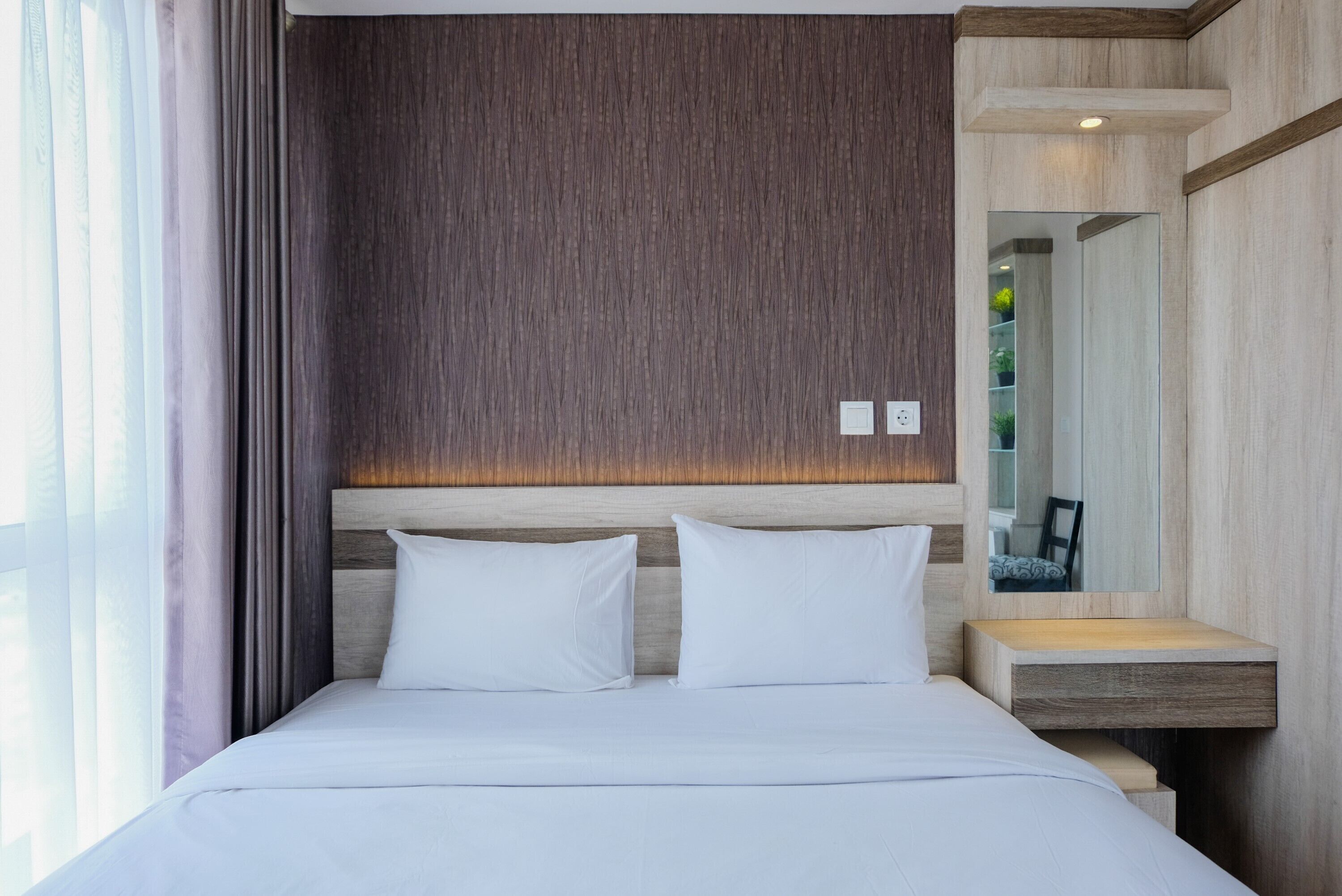 A modern hotel room with a wooden headboard, a bedside table, and a large mirror.