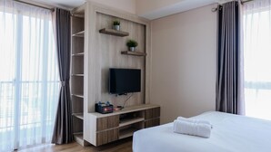 A hotel room with a bed, a TV mounted on a wooden wall, a shelf with plants, and a window with curtains. - Cozy and Luxurious Studio Casa de Parco Apartment (Pagedangan)