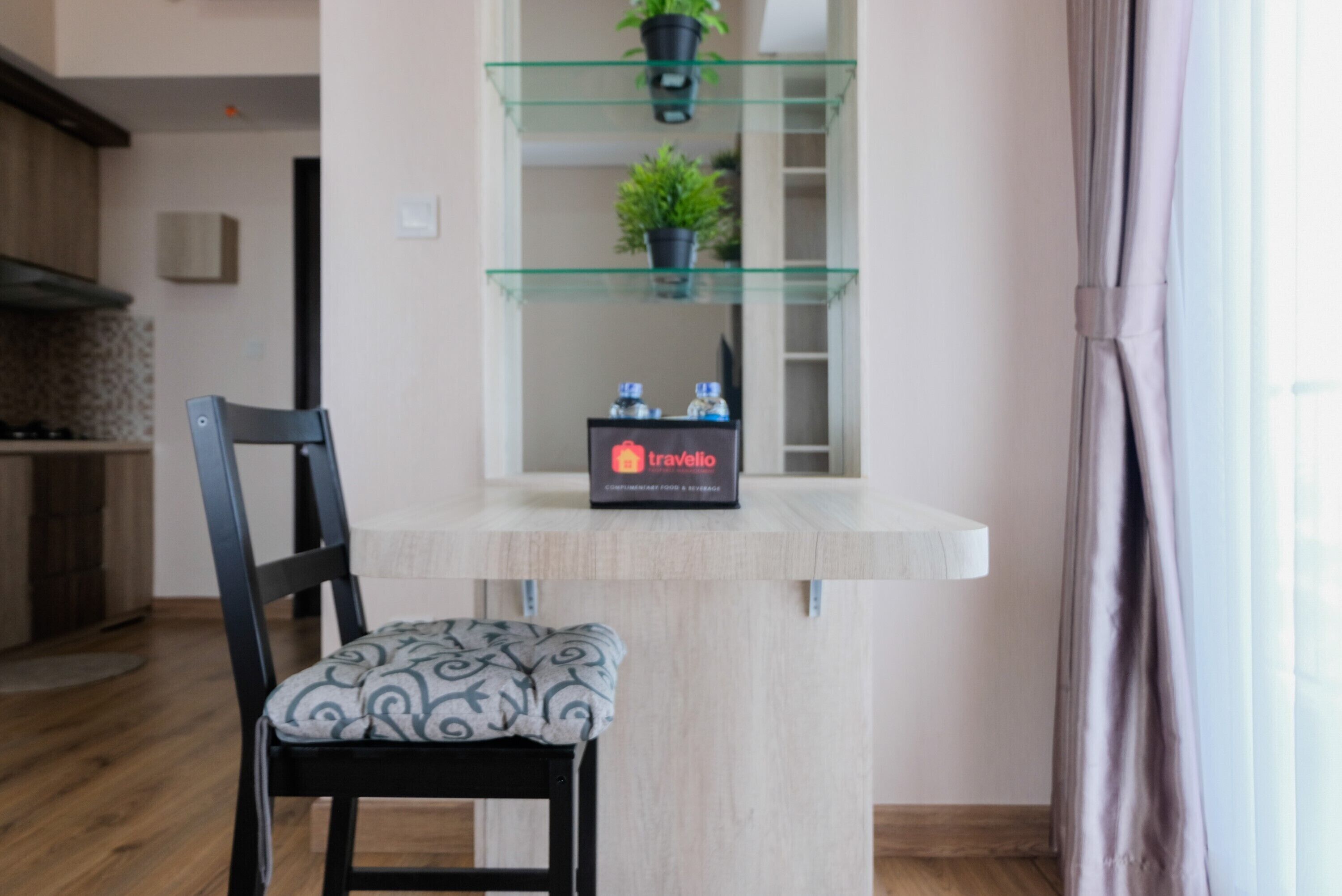 A modern kitchen with a wooden floor, a black chair with a patterned cushion, a glass shelf with plants, and a 'Travelio' sign on a white counter.