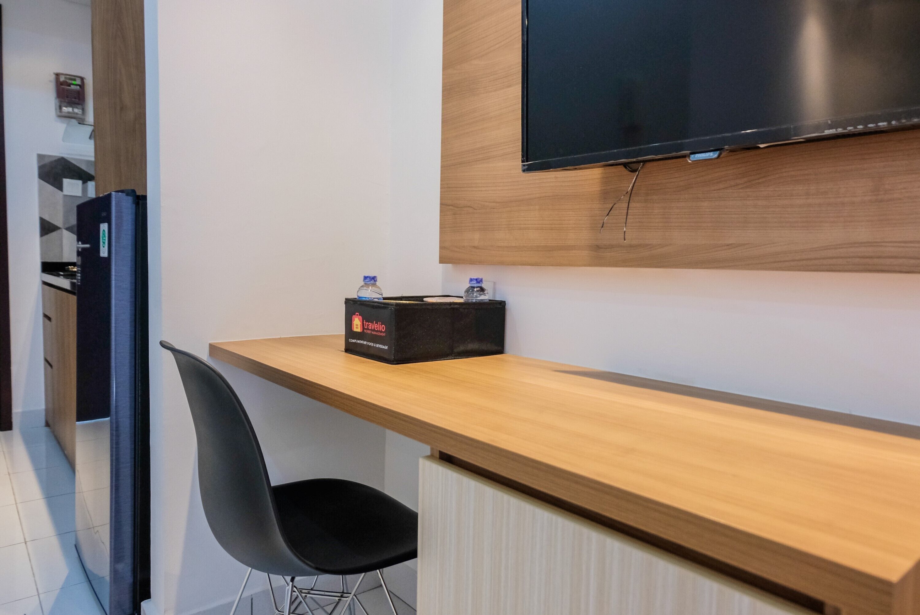 A modern hotel room with a wooden desk, a black chair, a flat-screen TV mounted on the wall, and a minibar.