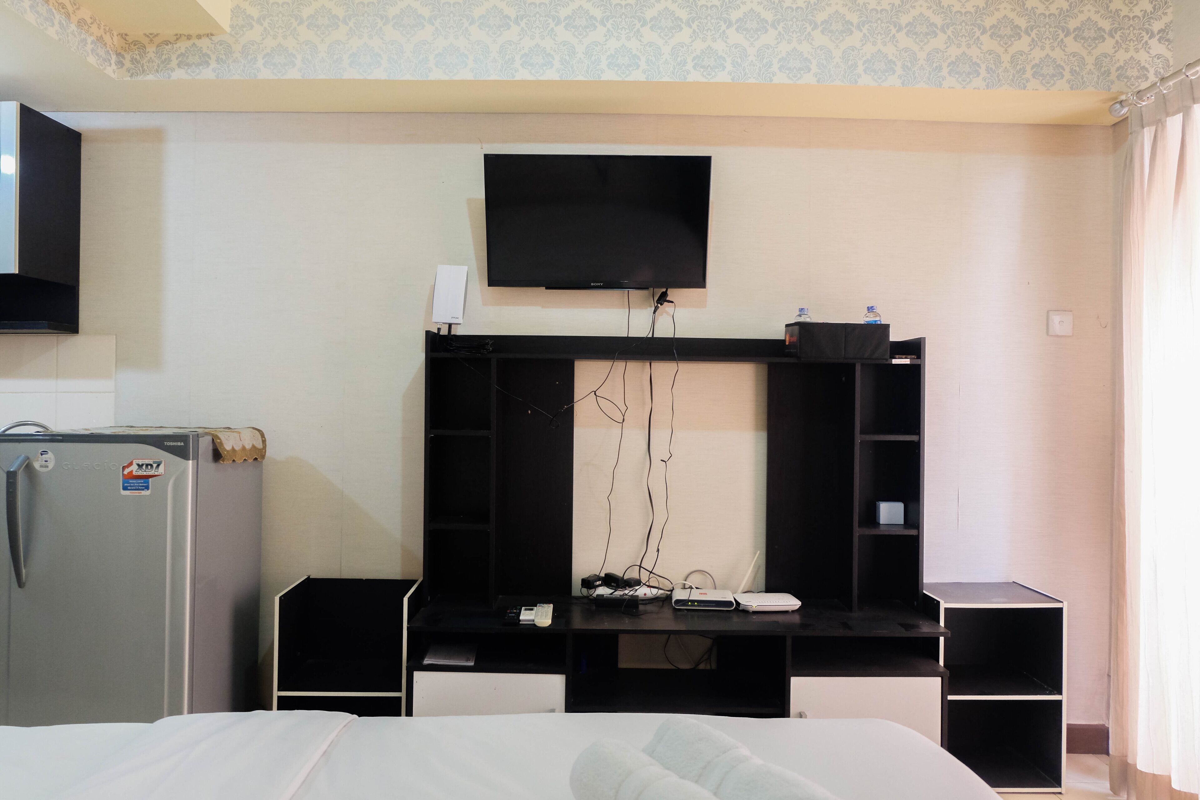 A hotel room with a bed, a desk with a flat-screen TV, a refrigerator, and a wardrobe.