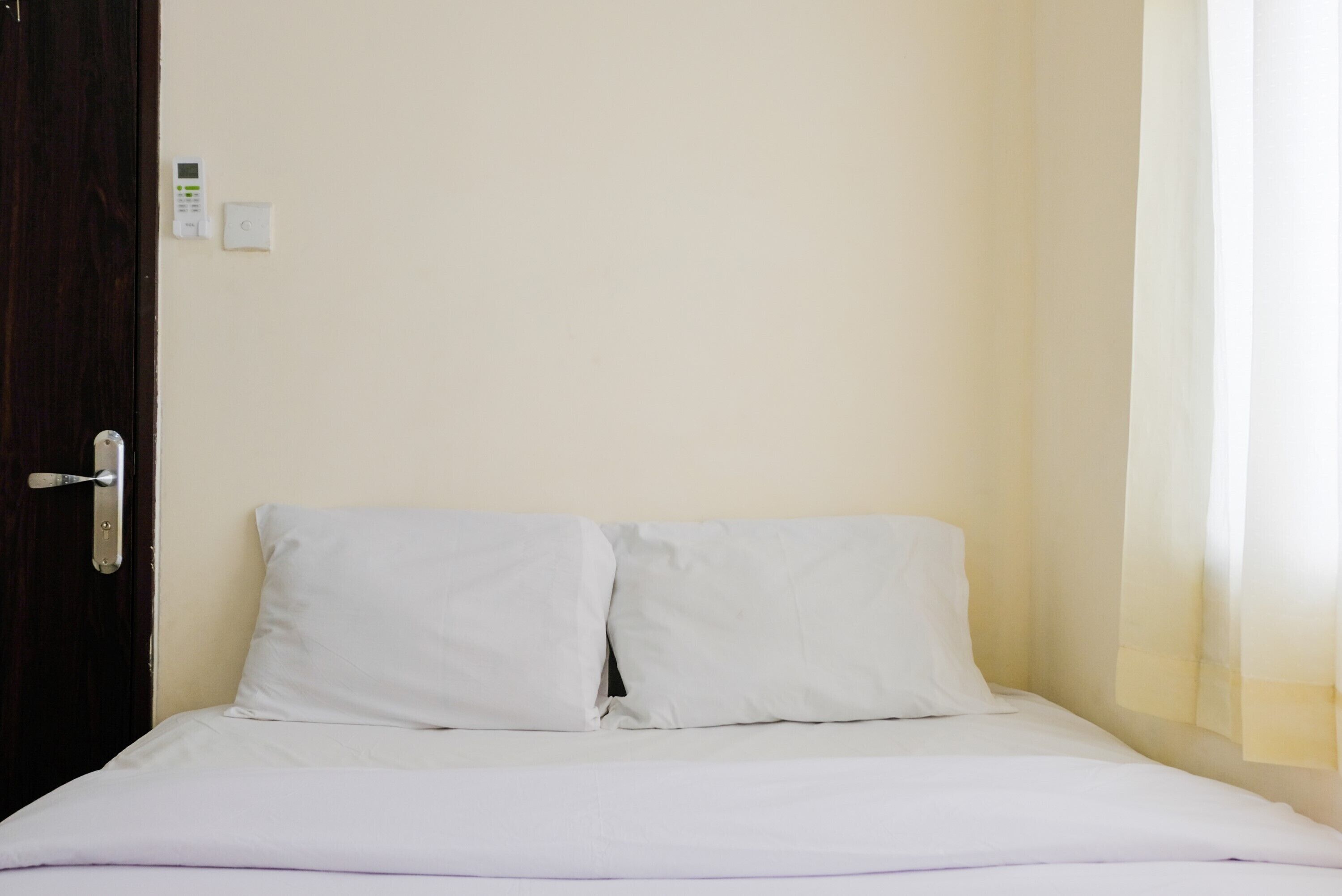 Een enkel bed met witte lakens, een houten deur en een lichtschakelaar aan de muur.