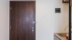 A closed wooden door with a keyhole and a handle, a small electrical outlet on the wall, and a kitchen area with a sink and countertop. - Highest Value Studio Room at Annora Living Apartment (Tangerang)