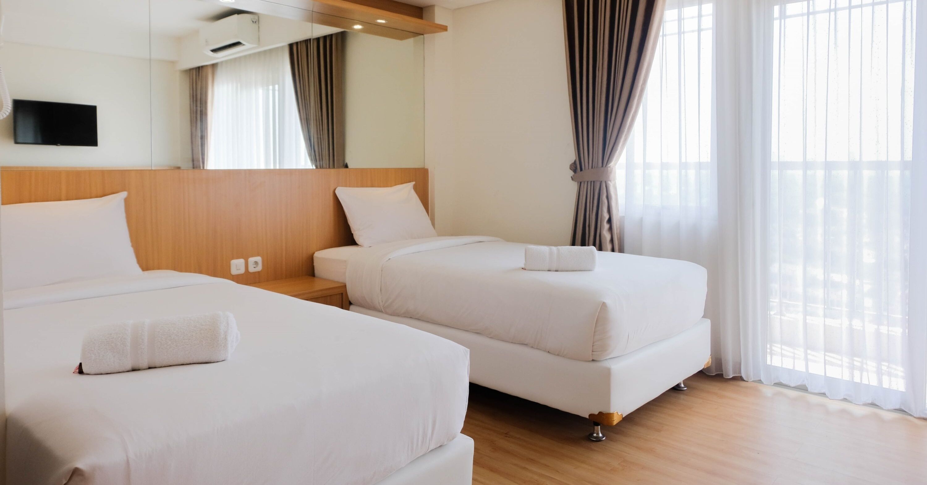 Ein Hotelzimmer mit zwei Betten, einem wandmontierten Flachbildfernseher und großen Fenstern mit Vorhängen.