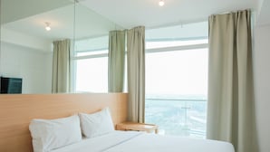 A hotel room with a large bed, a wooden headboard, a small wooden table, and a large window with curtains. - Cozy Studio Apartment at Tree Park City (Tangerang)
