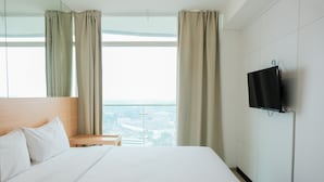A hotel room with a large bed, a television mounted on the wall, and a view of the city through large windows. - Cozy Studio Apartment at Tree Park City (Tangerang)