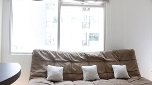 Ein beiges Sofa mit drei Kissen in Musteroptik, ein Fenster mit einem gemusterten Vorhang und ein Blick auf Gebäude im Freien.
