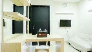 A room with a wooden desk, a white chair, a black door, a television mounted on the wall, and a bed covered with a white sheet.
