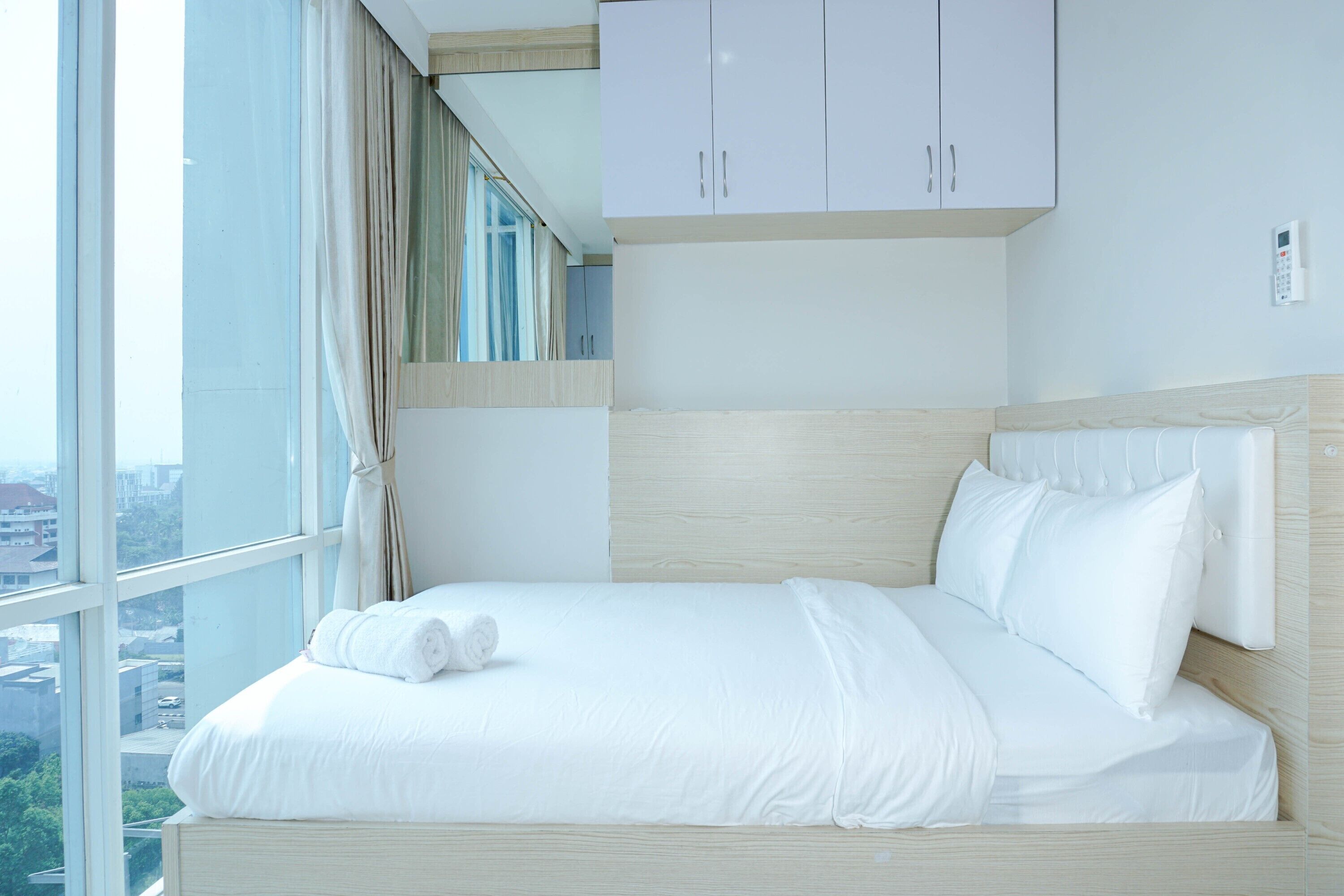 A hotel room with a large window, a bed with white linens, a wooden headboard, and a white wardrobe.