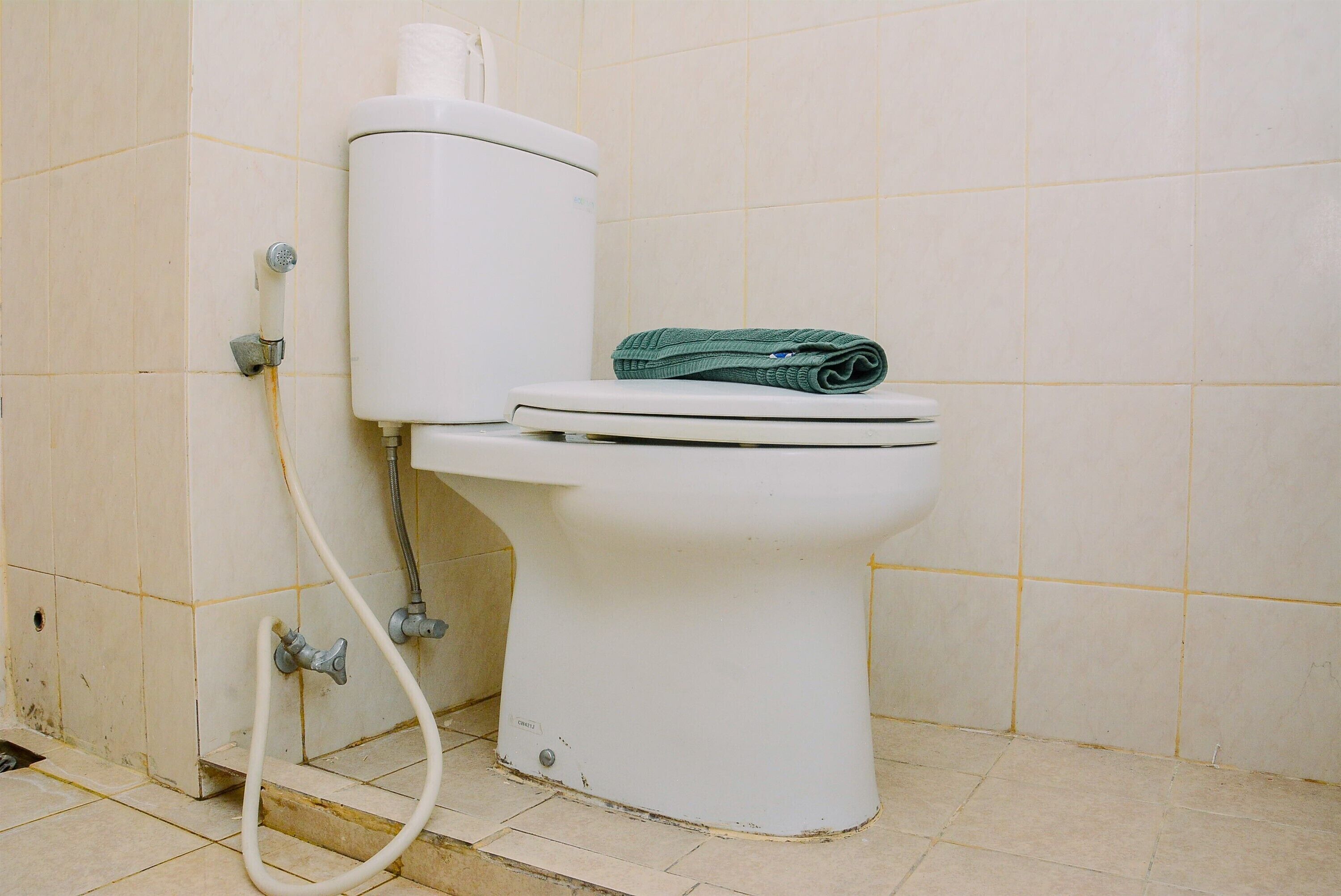 Inodoro con tanque y asiento blancos, una toalla verde sobre el asiento, y un tubo visible.