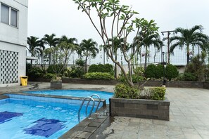 Una piscina con una escalera metálica, rodeada de palmeras y otra vegetación, con un edificio al fondo.