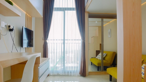 A modern living room with a flat-screen TV, a wooden desk, a white chair, and a yellow sofa. - Stylish and Convenient Studio Dave Apartment (Depok)