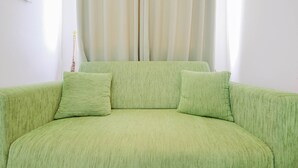Un canapé vert avec deux coussins, dans une pièce aux murs blancs et dotée d’une fenêtre avec des rideaux.
