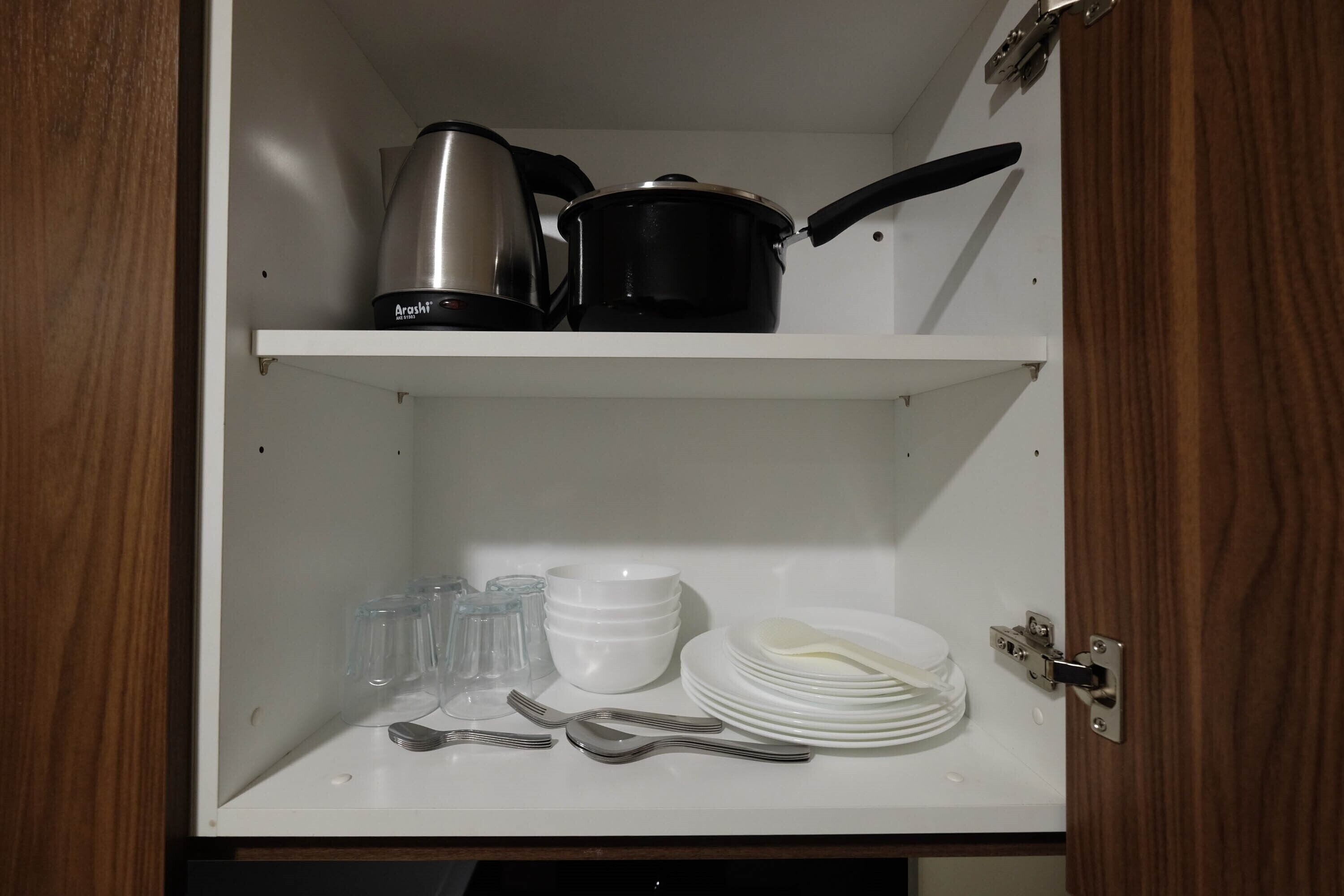 Een keukenkastje met een waterkoker, pan, glazen, kommen, borden en bestek.