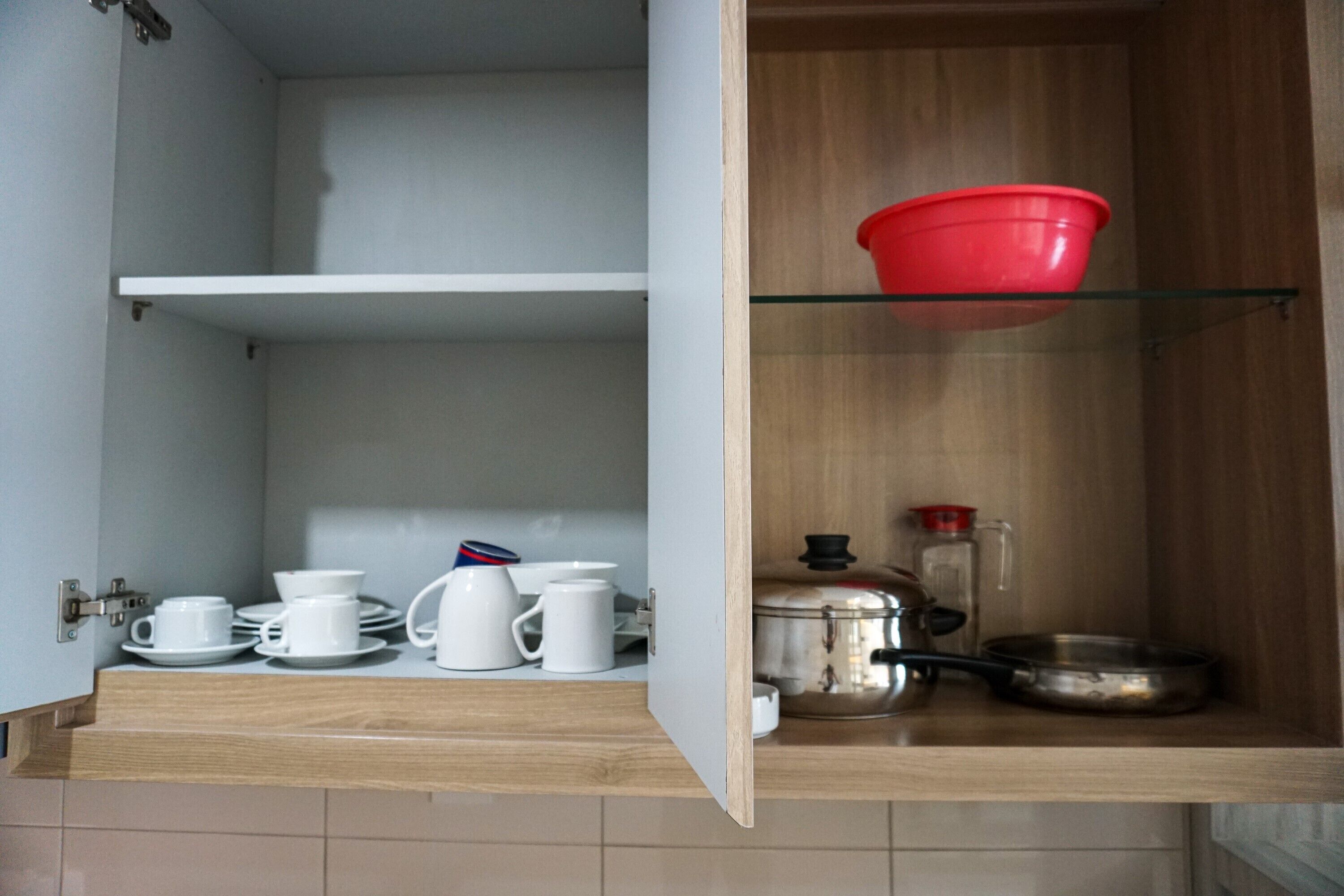 An open kitchen cabinet with white cups, saucers, a coffee pot, and a red bowl on a shelf.