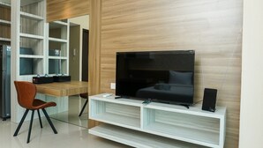 A modern living room with a flat-screen TV on a white entertainment unit, a wooden wall, and a built-in shelving unit. - Cozy Studio Apartment at Nine Residence connected to Mall (Jakarta)