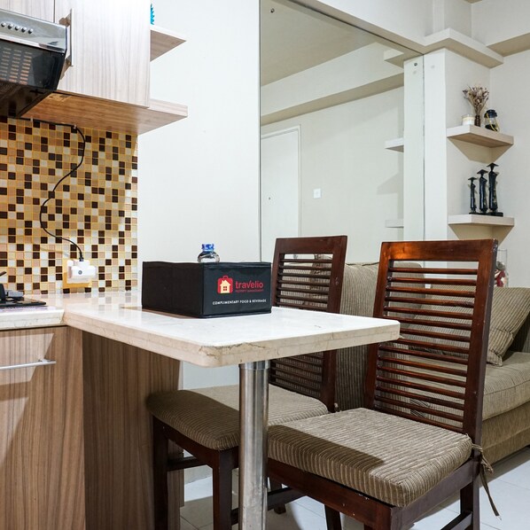 A compact kitchen with a tiled backsplash, a countertop, and a sink. There is a black box labeled 'Travelio Property Management' on the counter. Two wooden chairs with cushioned seats are placed at the counter.