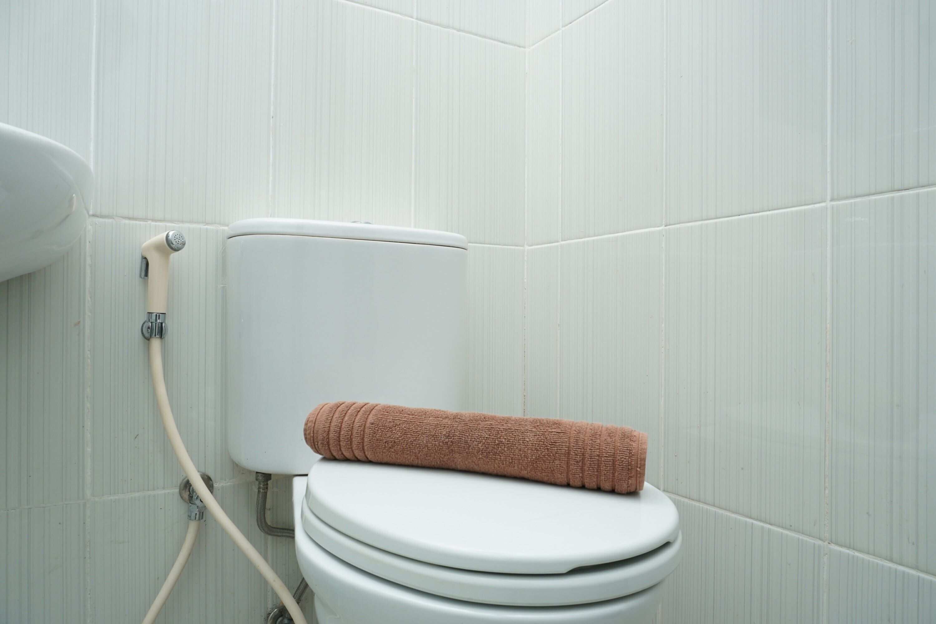 A white toilet with a beige towel on top, a shower hose, and a white tiled wall.