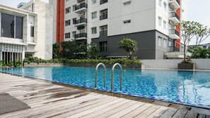 Une piscine entourée d’une terrasse en bois, elle-même bordée de bâtiments modernes dotés de balcons et de verdure.