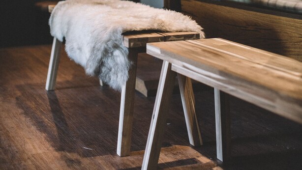 Un banco de madera con un plaid de piel, una pequeña mesa con un jarrón de flores, y una zona de bar al fondo.