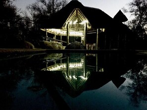 A building with a glass facade reflecting on a body of water, surrounded by trees. - River Rock Guesthouse (Ngwathe)
