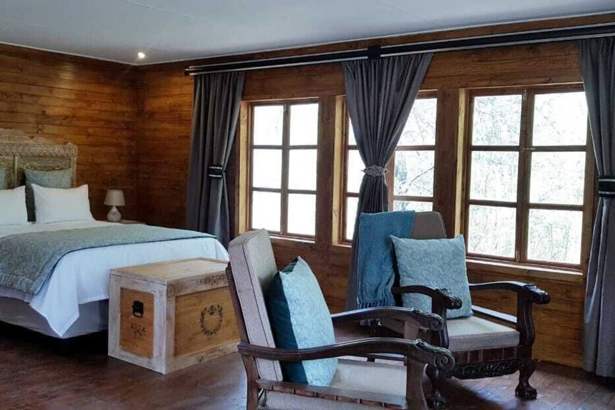 A wooden cabin-style bedroom with a large bed, two bedside tables, a chair, and a window with curtains.