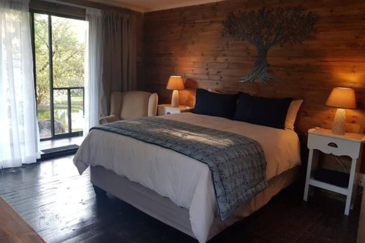 A bedroom with a large bed, two bedside tables with lamps, a wooden wall with a tree mural, and a sliding glass door leading to a balcony.