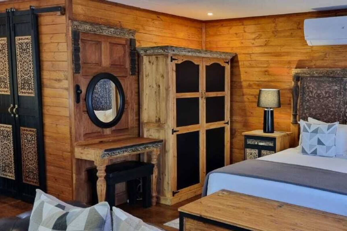 A wooden-themed bedroom with a large bed, two bedside tables with lamps, a wooden wardrobe, and a small desk with a mirror.