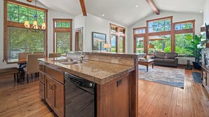 Una cocina amplia con encimera de granito, armarios de madera y lavavajillas. Cuenta con ventanas grandes con persianas, un comedor con sillas y una sala de estar con sofá y mesa de centro.
