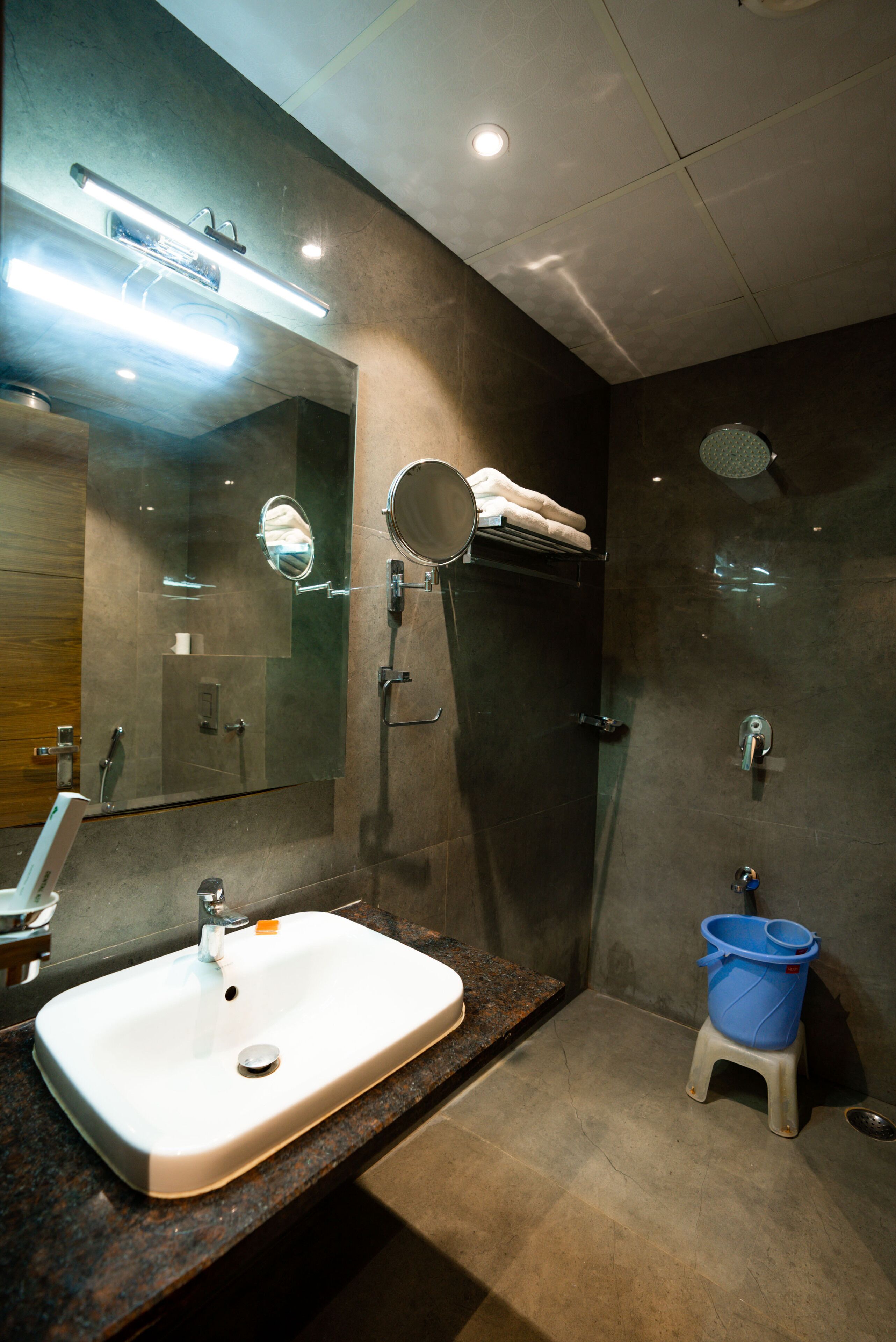 A modern bathroom with a large mirror, a sink, a shower, and a blue bucket.