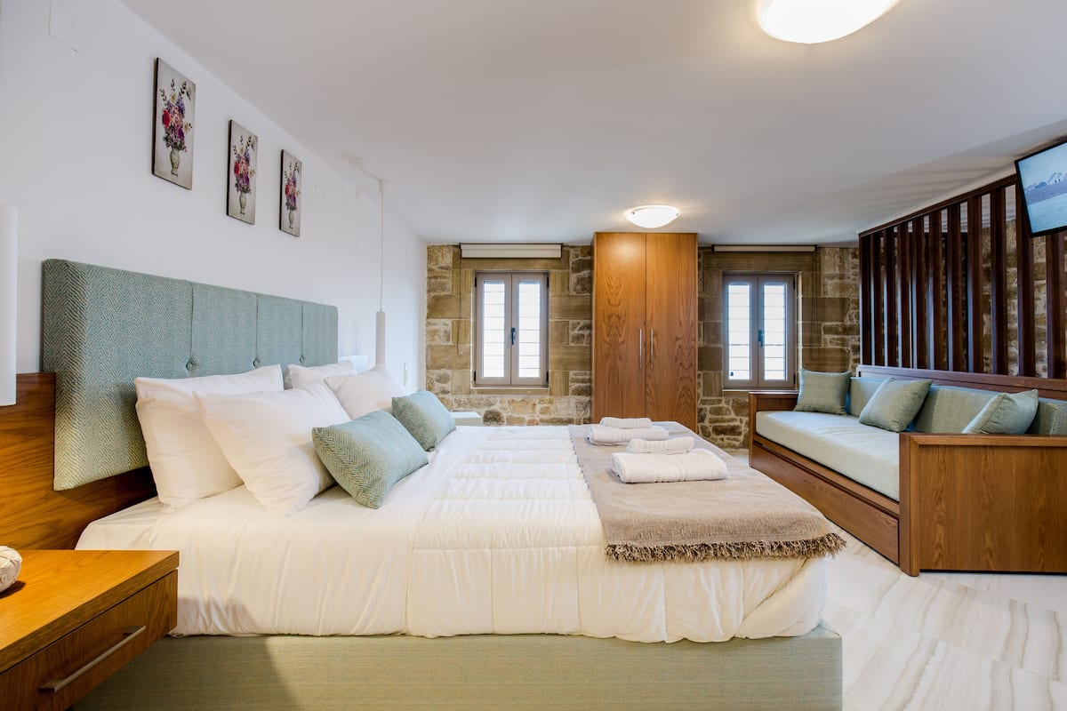 A bedroom with a large bed, a wooden bench, stone walls, and a TV on the wall.