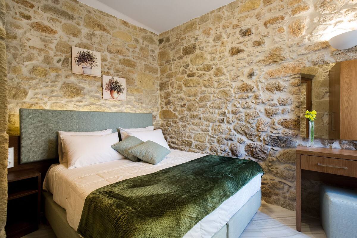 A bedroom with a stone wall, a bed with white linens, a green throw blanket, a nightstand, and a wooden dresser.