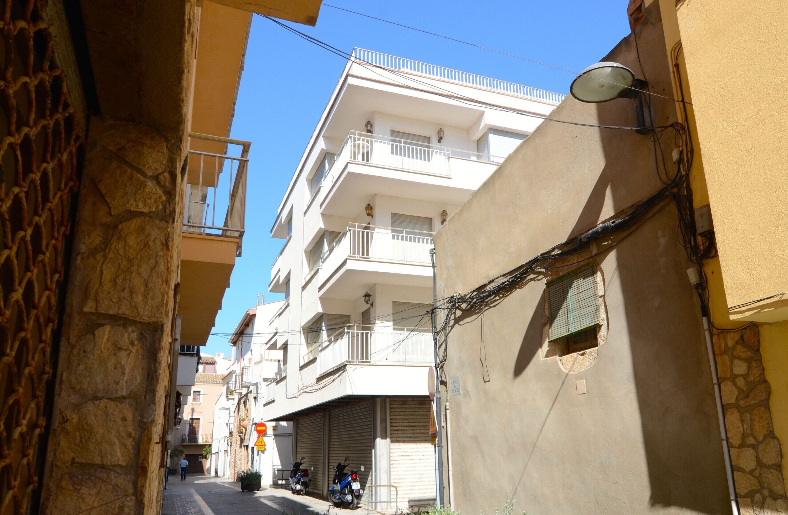 A narrow street with buildings on either side, featuring balconies and a clear blue sky.