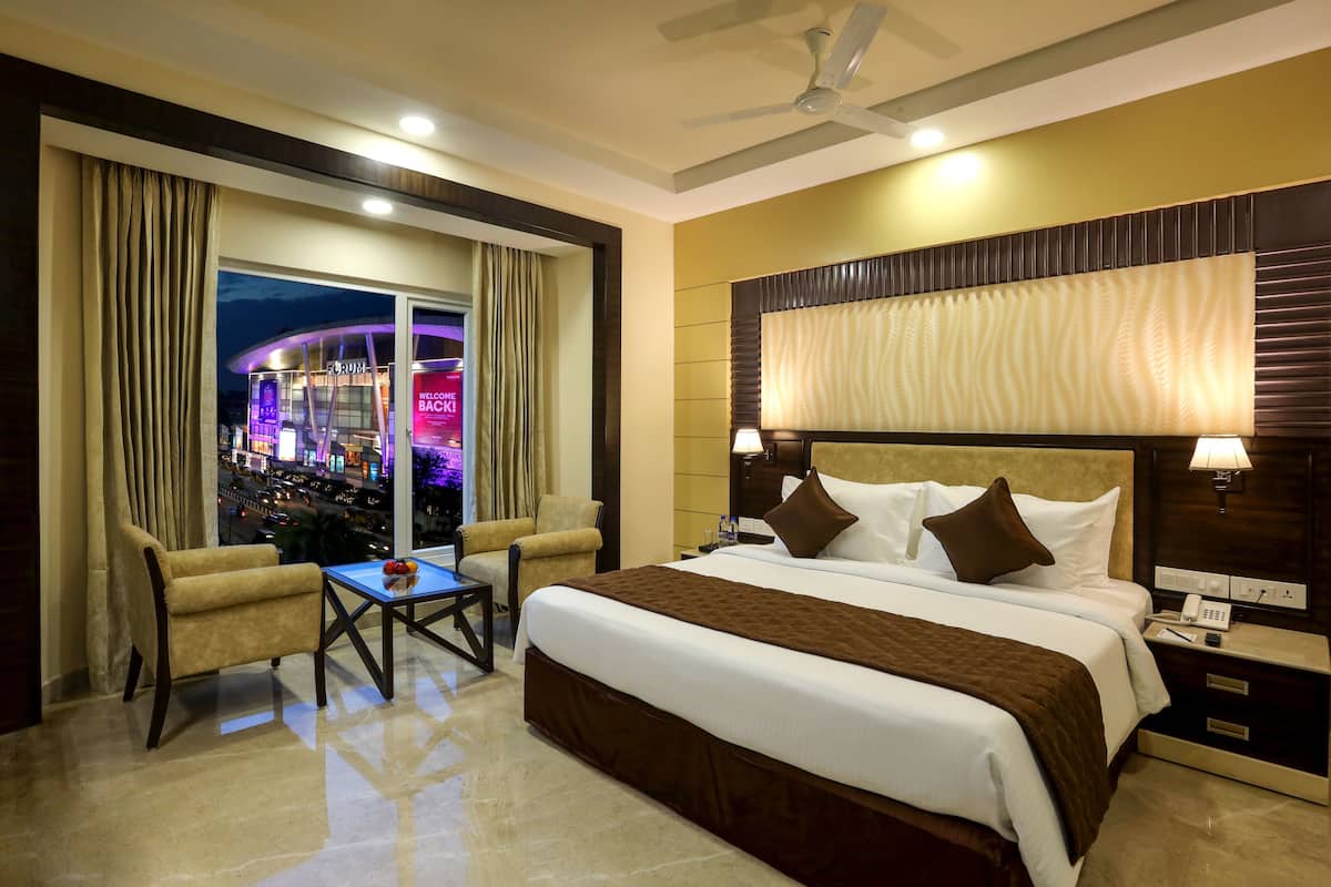 A hotel room with a large bed, a desk, two armchairs, a view of a cityscape, and a ceiling fan.