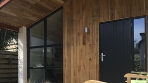 A wooden building with a large glass door and a black door, a wooden bench, and a flower bed with yellow flowers. - Spacious house & spa near forest, beach and sea and the cozy village of Bergen (Bergen)
