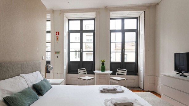 Een hotelkamer met een bed, twee stoelen, een klein tafeltje, een televisie en grote ramen.