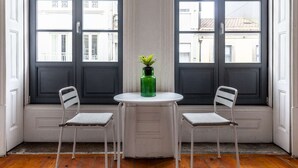 Een kamer met twee witte metalen stoelen en een kleine witte tafel, een groene vaas, en een raam met uitzicht op gebouwen.