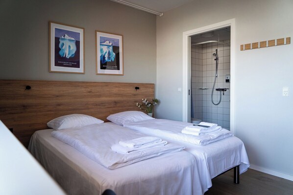 A hotel room with two beds, a wooden headboard, a shower area, and two framed pictures on the wall.