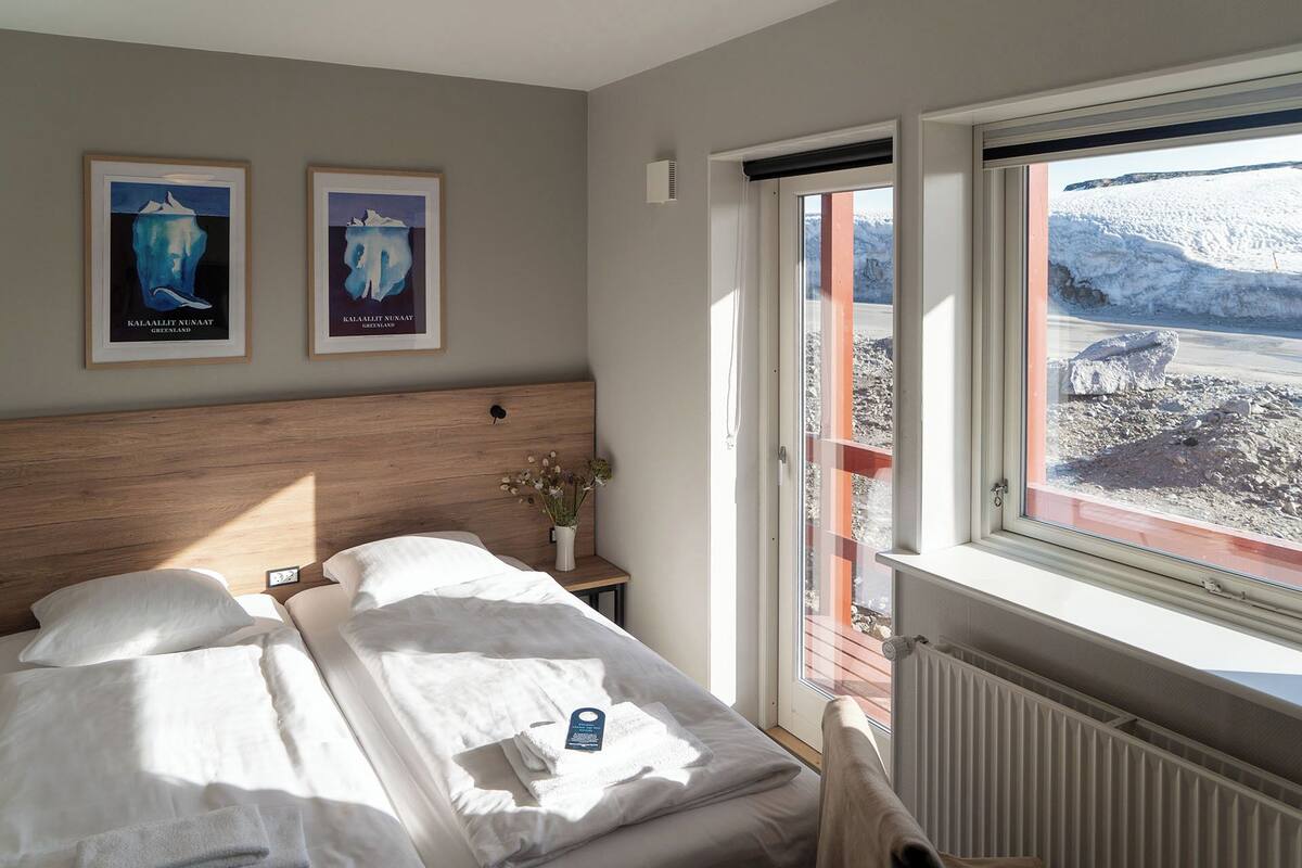 A bedroom with a wooden headboard, a bed with white linens, a nightstand with a vase of flowers, and a window with a view of a snowy landscape.
