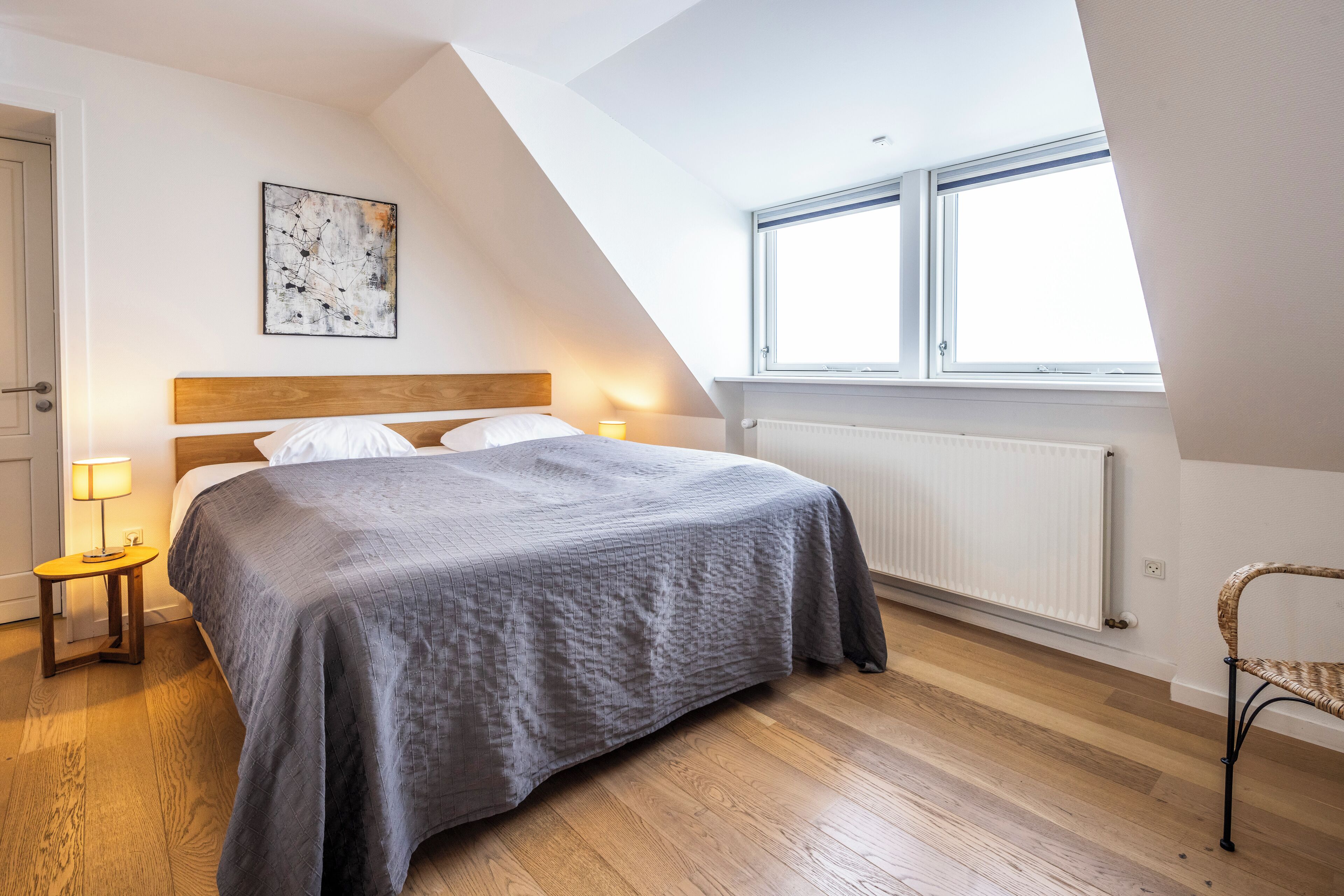 A bedroom with a bed, a nightstand, a chair, a window, and a radiator.