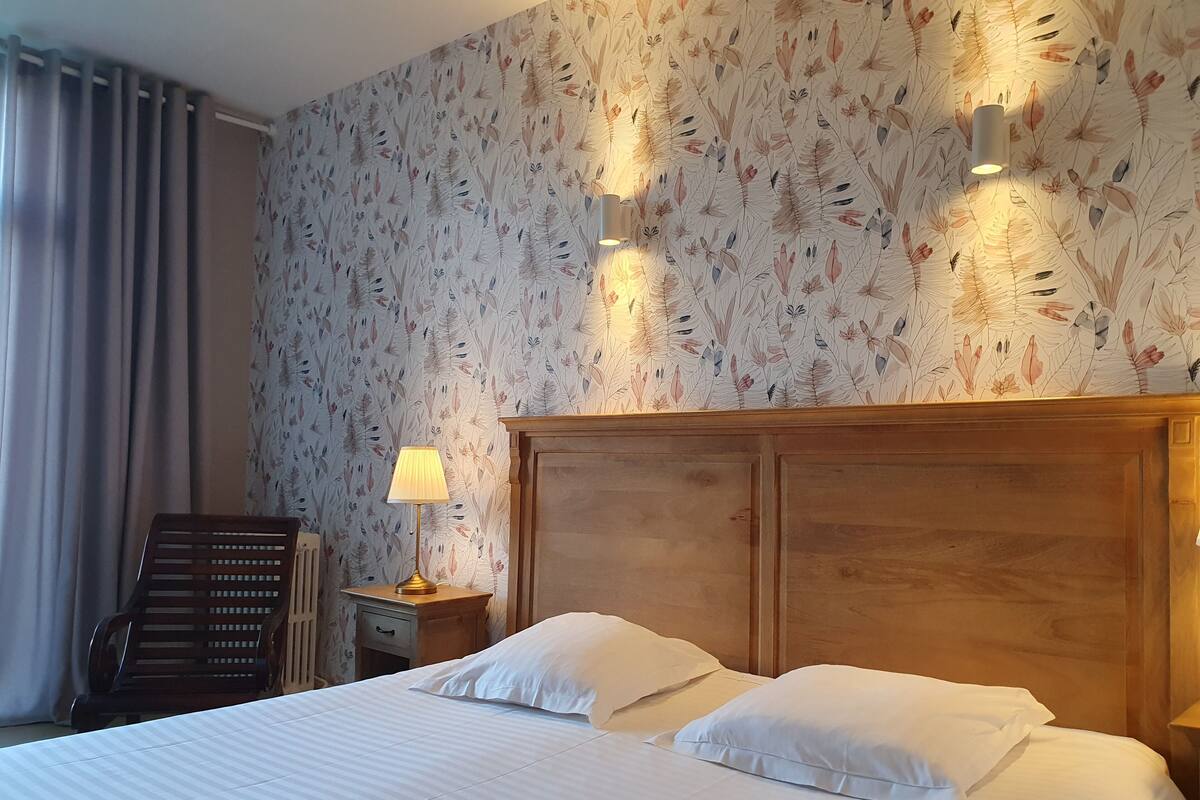 Een hotelkamer met een groot bed, een houten hoofdeinde, een nachtkastje met een lamp, een stoel en een radiator.