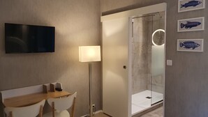 Ein modernes Zimmer mit Flachbildfernseher, Schreibtisch mit Stühlen, Stehlampe und einer Duschkabine aus Glas.
